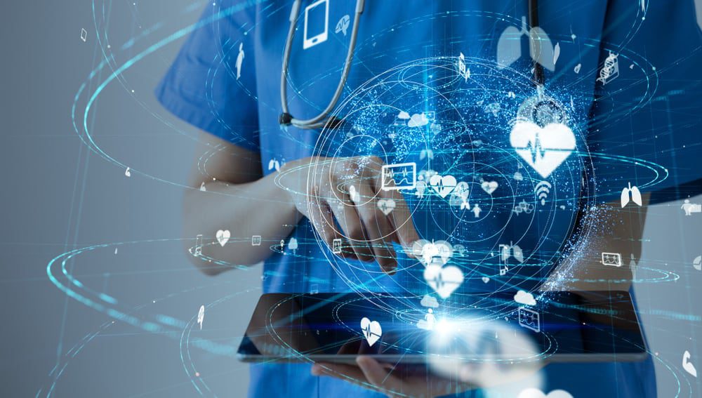 Healthcare network design is visualized as a bunch of glowing healthcare technology icons above a tablet being used by a person wearing scrubs
